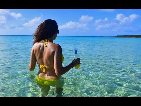 Back view of a person in a yellow bikini waist-deep in clear turquoise water, holding a blue-and-white bottle, sunny beach scene.