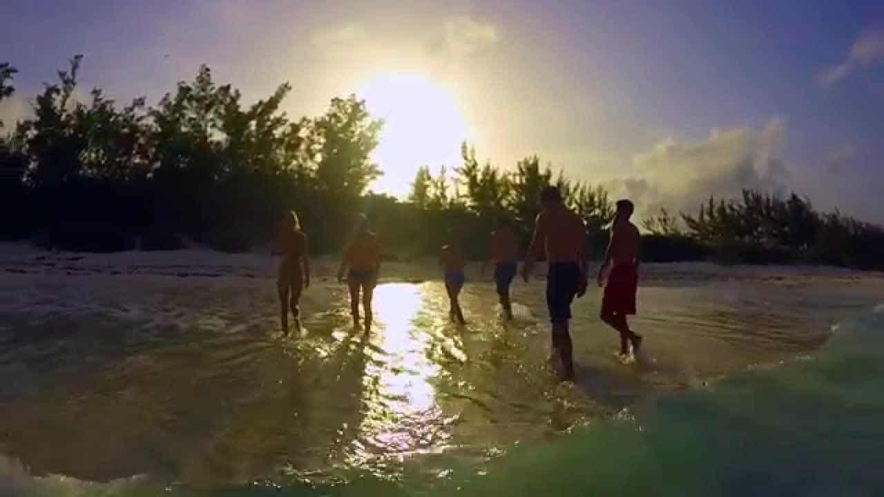 Group of people wading through shallow water on a sandy beach at sunset, with trees silhouetted in the background.