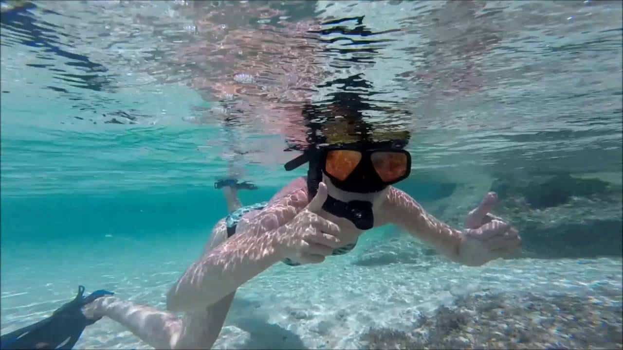 Snorkeler underwater giving thumbs up, wearing a mask and snorkel in clear tropical water with a rocky seabed beneath.