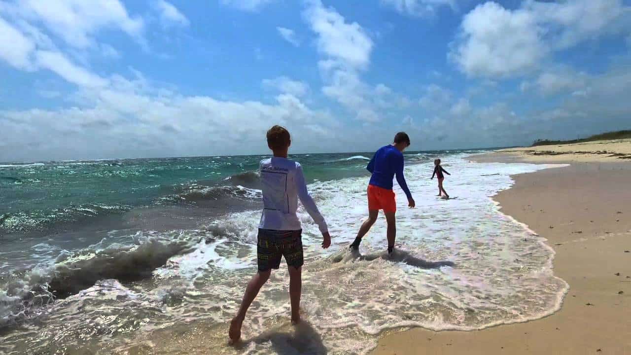 Three people walk along a sunny beach near the surf, turquoise water and white foam washing onto the sand.
