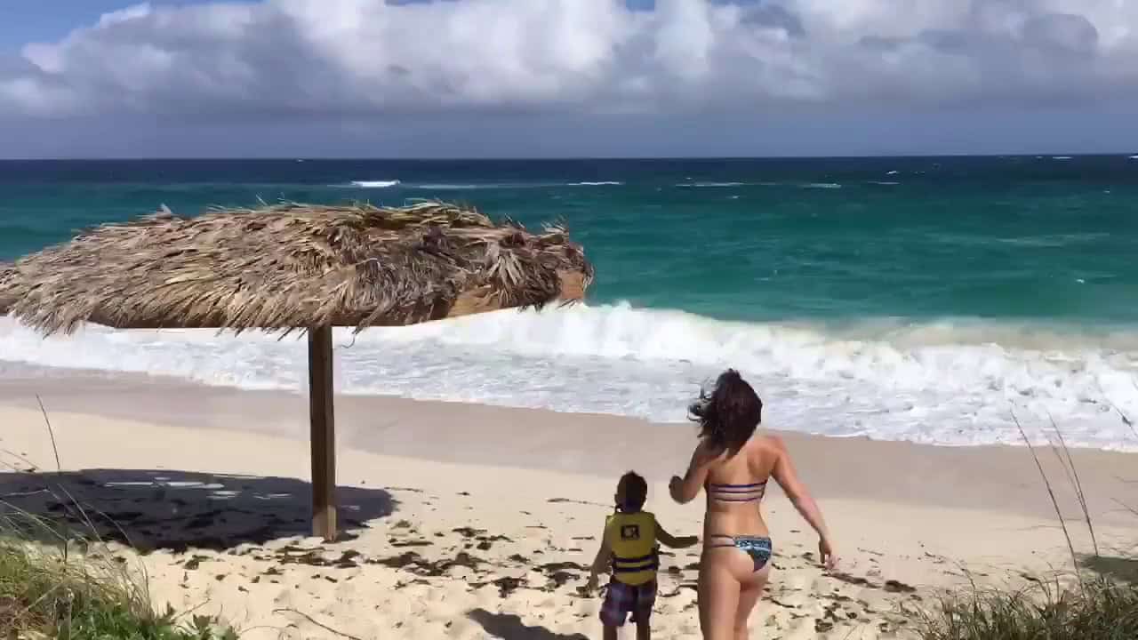 A woman in a bikini and a child in a life jacket walk toward the turquoise ocean on a sandy beach, near a thatched umbrella.