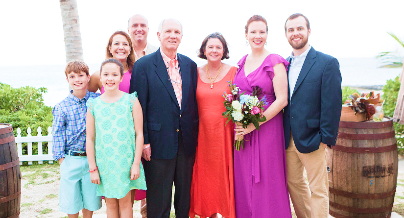 celebrate-togethe-your-eay - Button Wood Reserve - Resorts In Eleuthera Bahamas Group portrait of nine people at a sunny beach wedding, with the bride in a purple dress holding a bouquet and family members in bright outfits, ocean in the background.